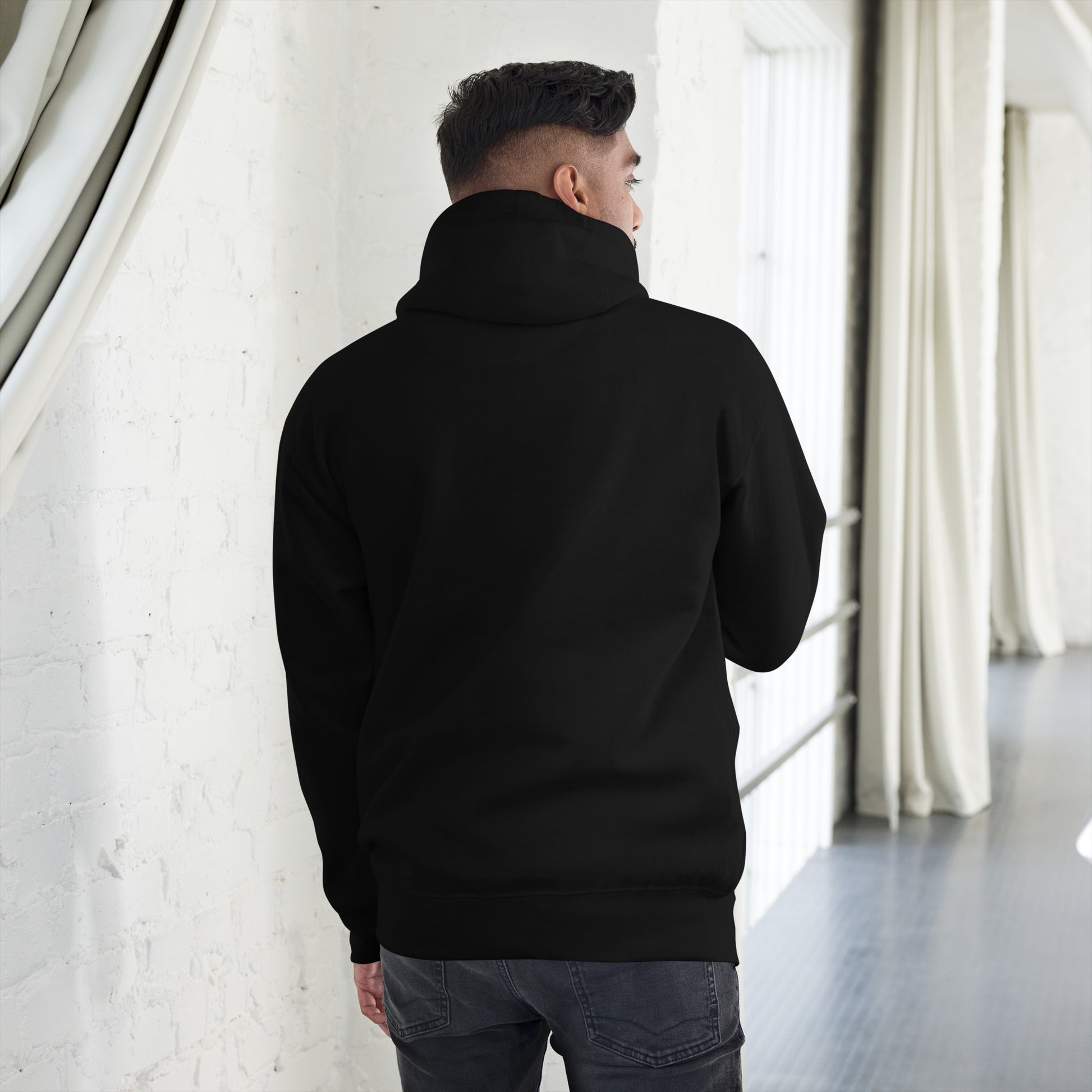 Person facing backward, showing the back of a black hoodie standing in a room with white curtains and a white wall.